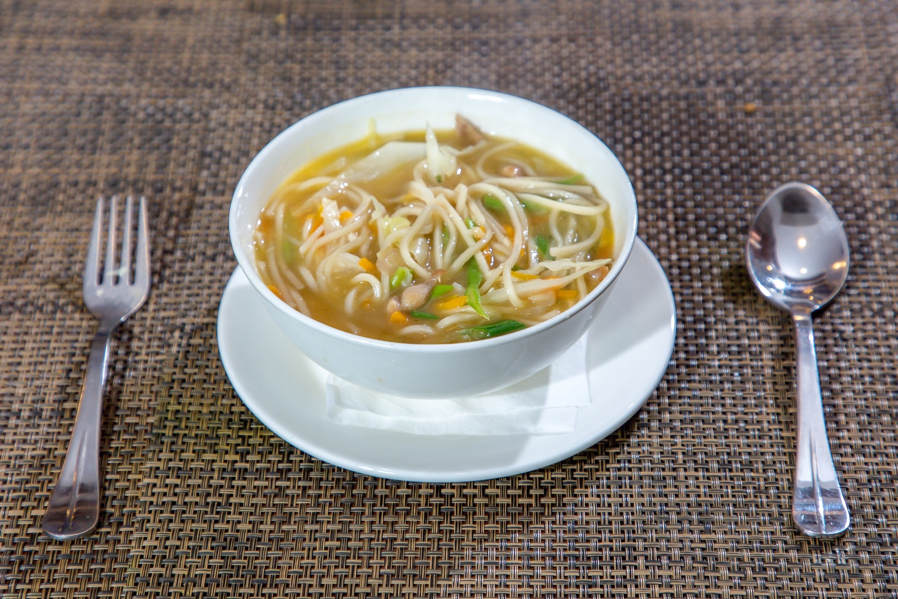 Un tazón de sopa de fideos sobre un plato blanco, con cuchara y tenedor sobre un mantel tejido.