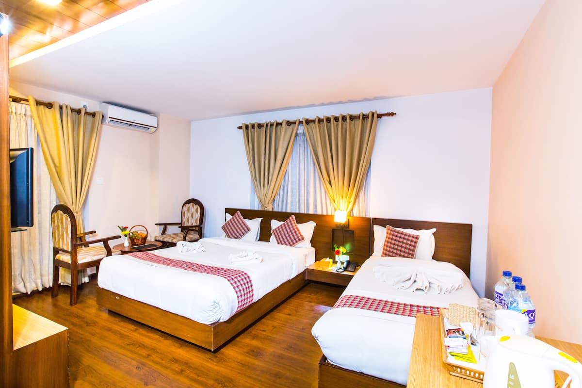 A hotel room with two beds, wooden flooring, a desk with a chair, a nightstand, and an air conditioning unit.