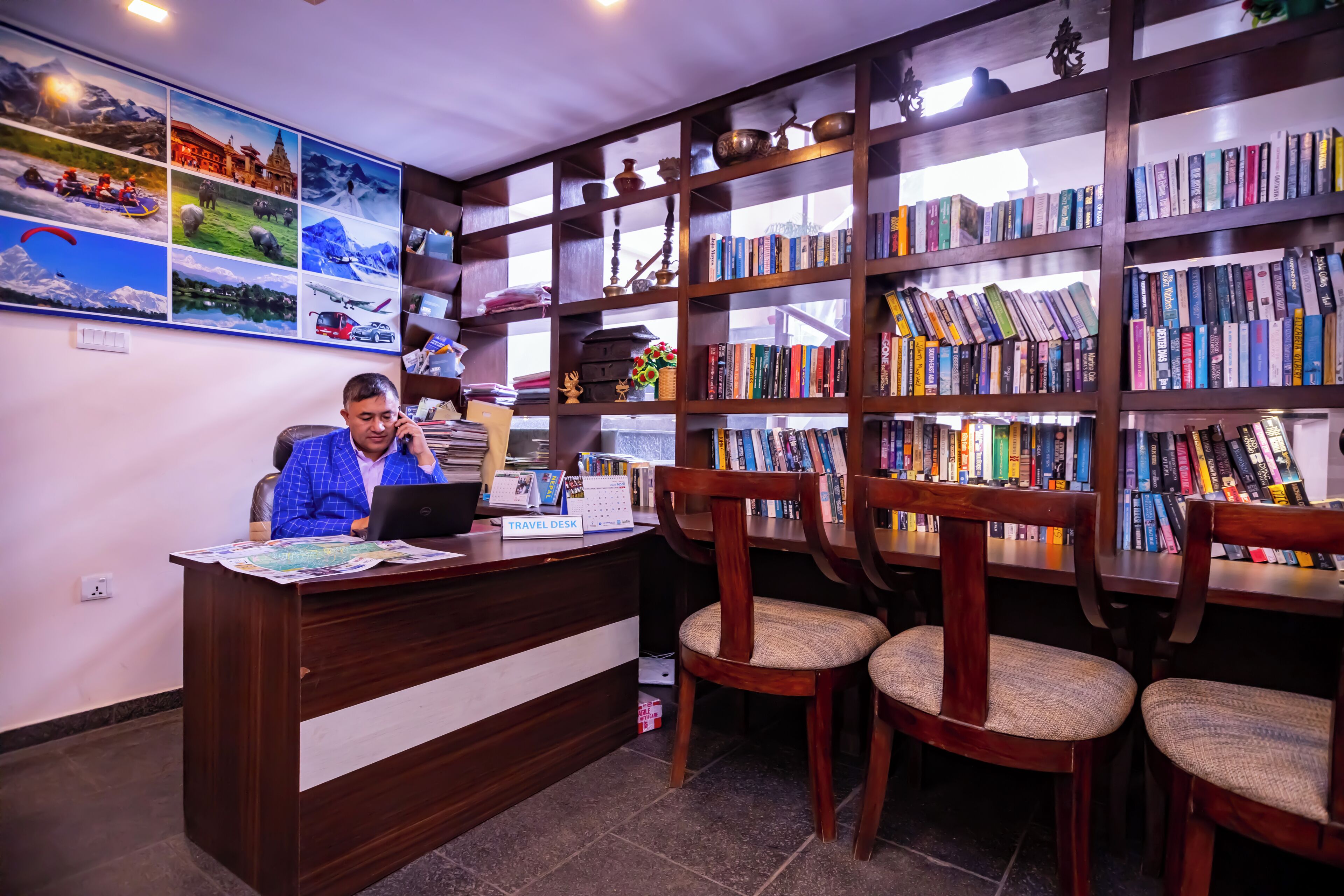 A reception desk with a laptop, a travel desk sign, and a wall with framed pictures. There are wooden bookshelves filled with books and a wooden chair with a cushion.