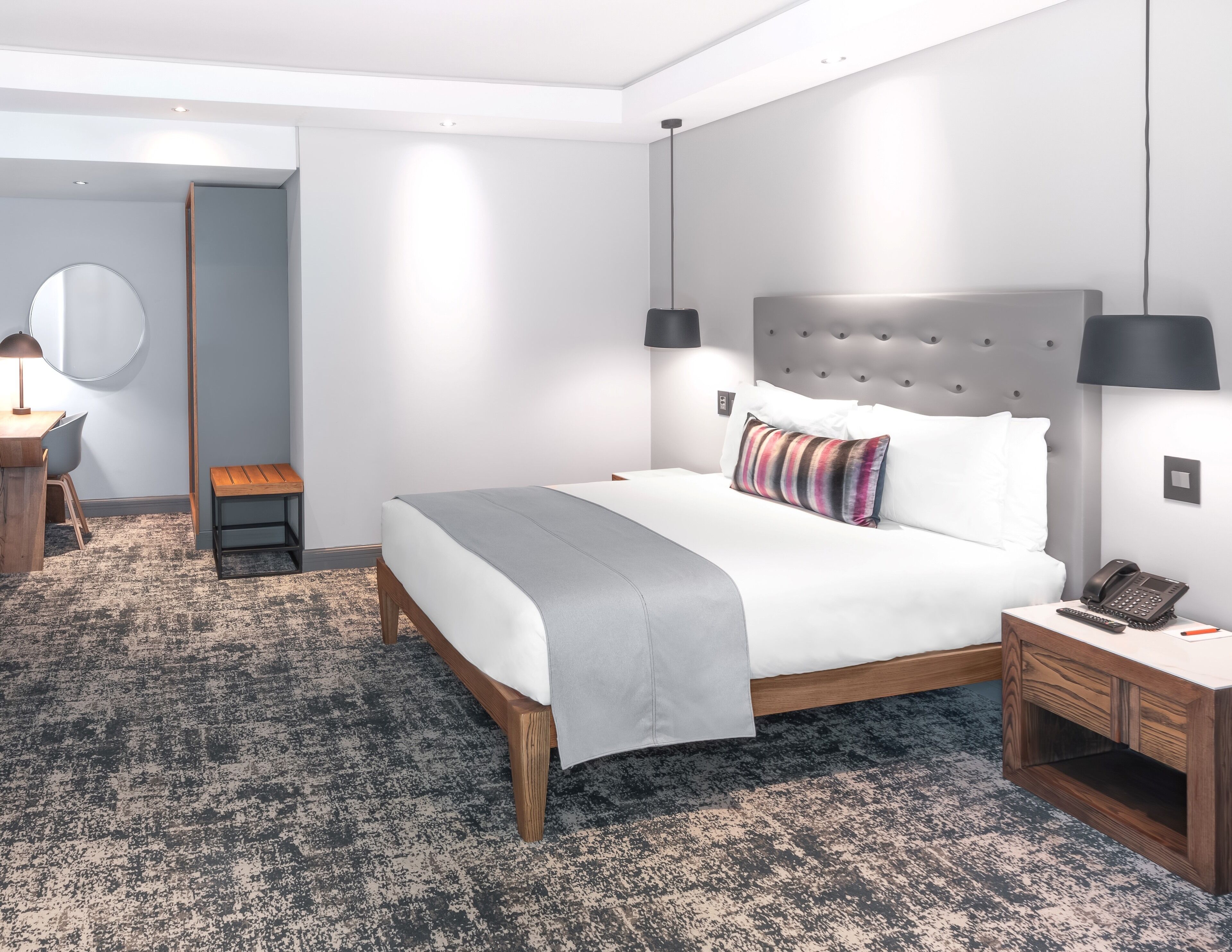 A modern hotel room with a large bed, a bedside table with a phone, a small stool, a desk with a chair, and a round mirror on the wall.