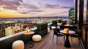 Una terraza en la azotea con mesas y sillas, vistas de la ciudad y un atardecer.
