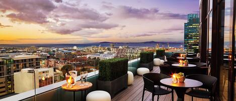 A rooftop terrace with tables and chairs, a view of the cityscape, and a sunset.