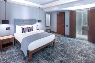 A modern hotel room with a large bed, a bedside table, a nightstand, a wardrobe, and a bathroom visible through a sliding door.