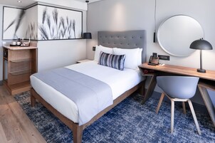 A modern bedroom with a large bed, bedside table, desk, chair, and a round mirror.