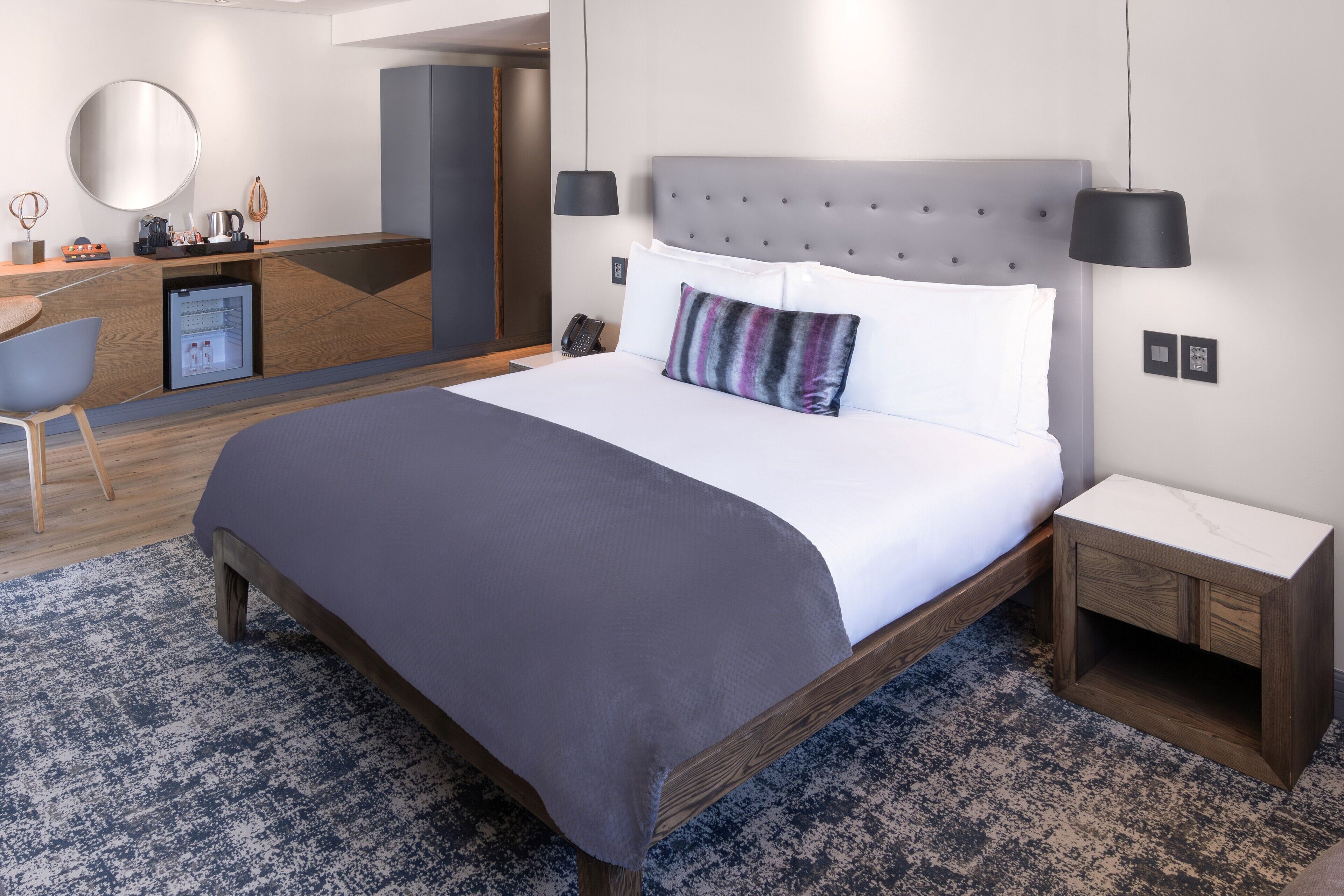 A modern hotel room with a large bed, a desk, a mirror, and a small refrigerator.