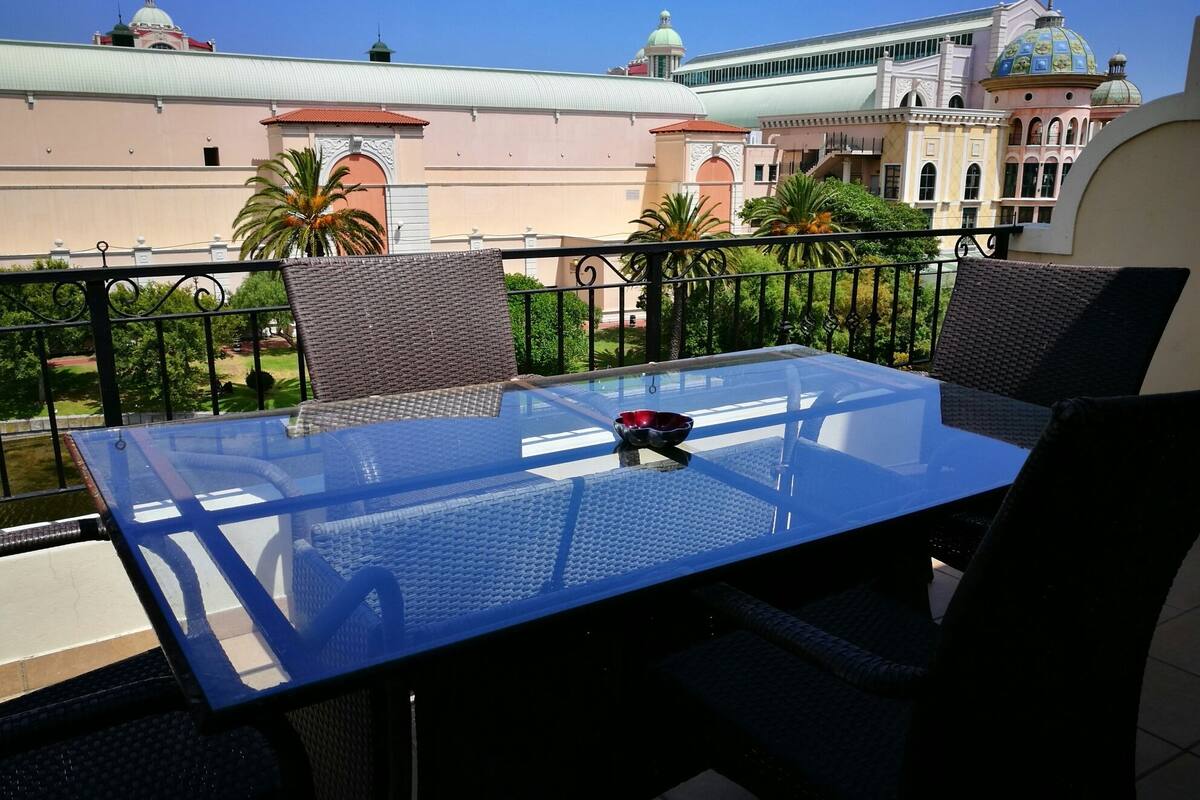 A balcony with a glass table and wicker chairs, overlooking a building with domed roofs and arched windows.
