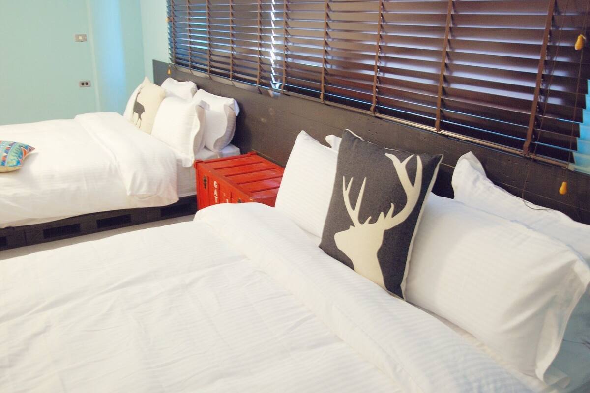 A hotel room with two beds, one with white bedding and the other with a patterned pillow. There is a red crate between the beds, and the room has a window with blinds.