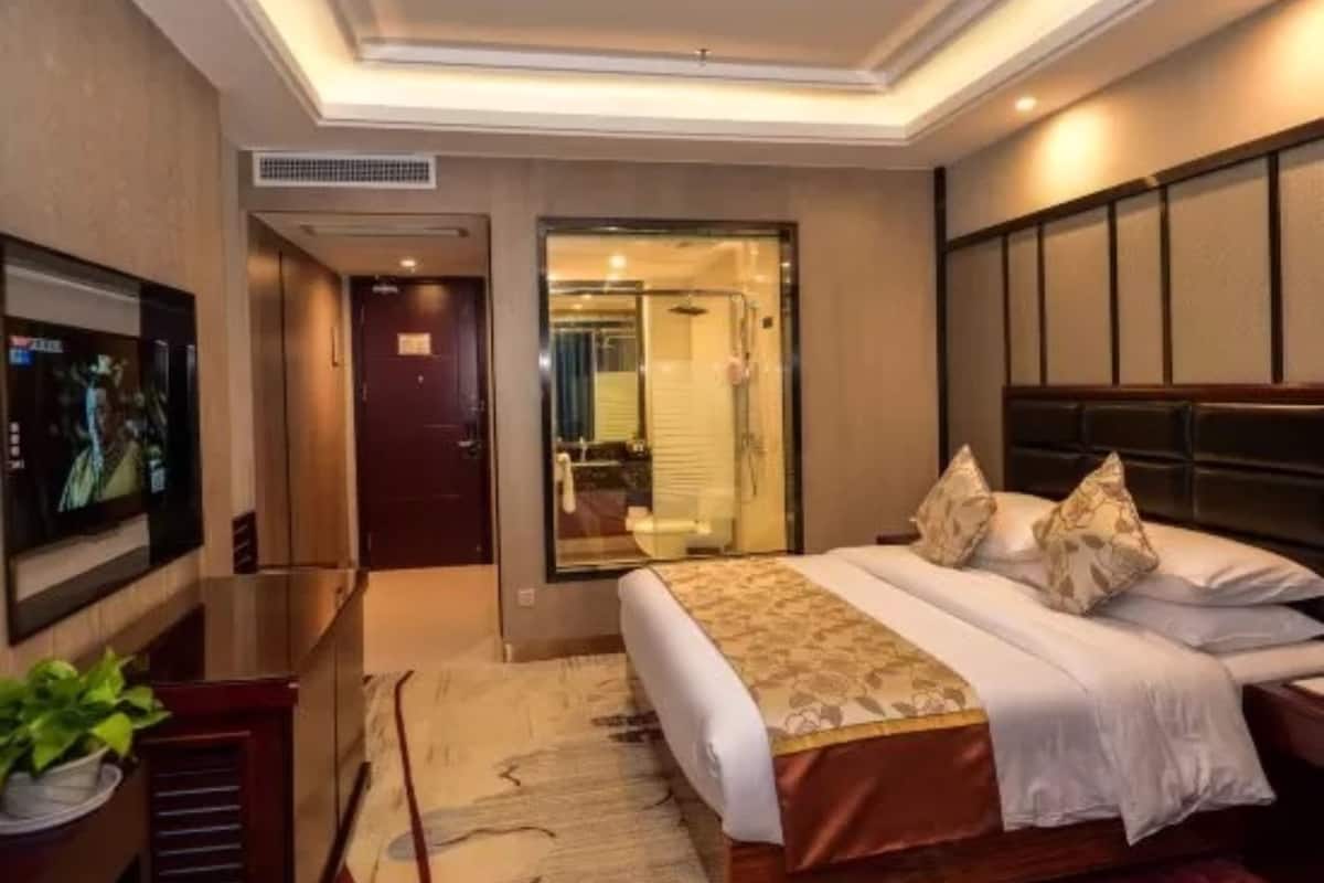 A hotel room with a bed, a TV, a desk, and a bathroom visible through a glass door.