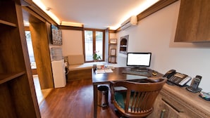 A wooden desk with a computer, phone, and various office supplies. There is a chair with a green cushion, a small trash can, and a water dispenser. The room has wooden flooring and a window with a view of the outdoors. - Kumluk Konak Hotel (Istanbul)
