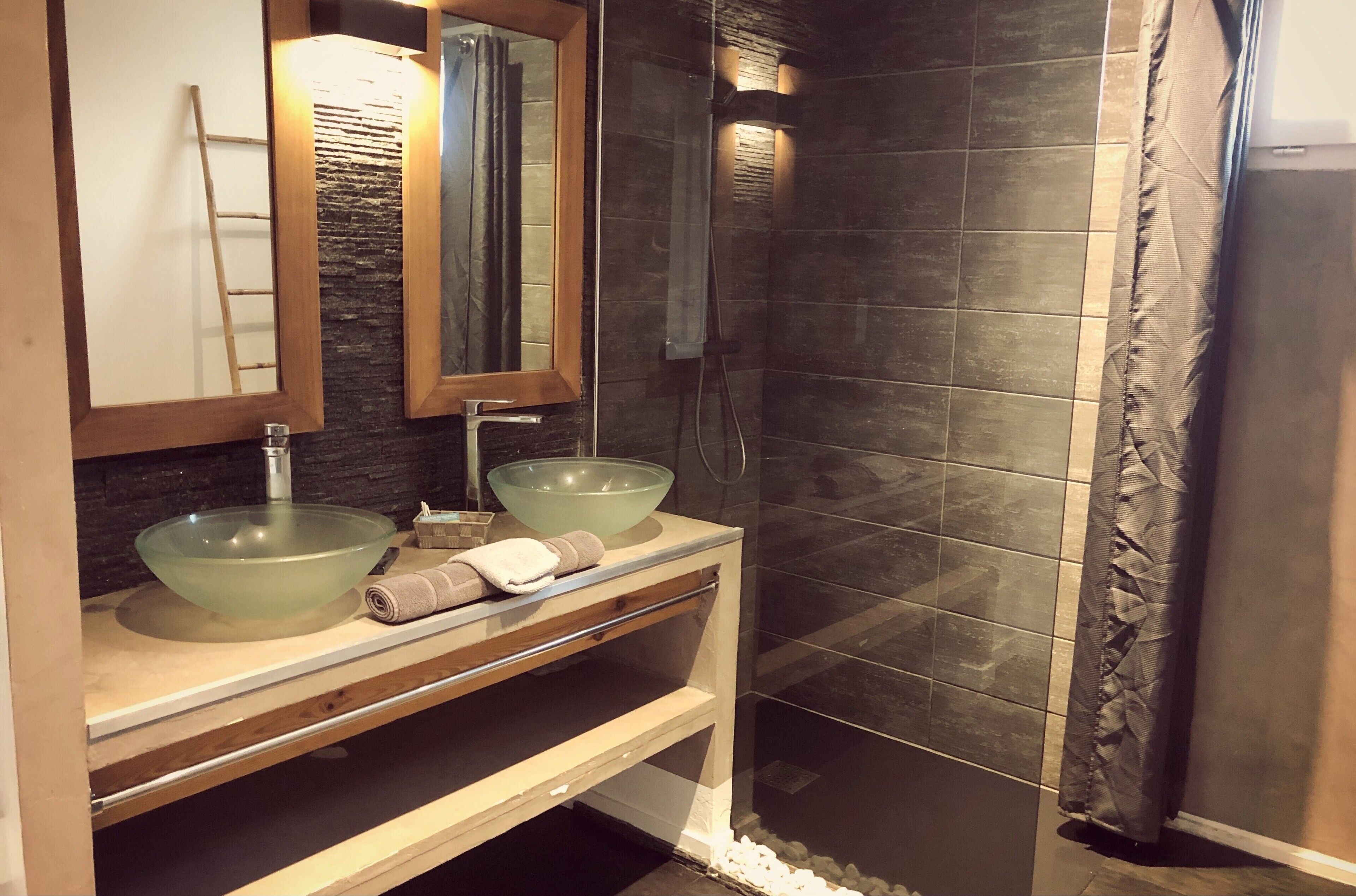 Une salle de bain avec un lavabo en verre, un meuble-lavabo en bois, un miroir et une douche aux carreaux foncés.