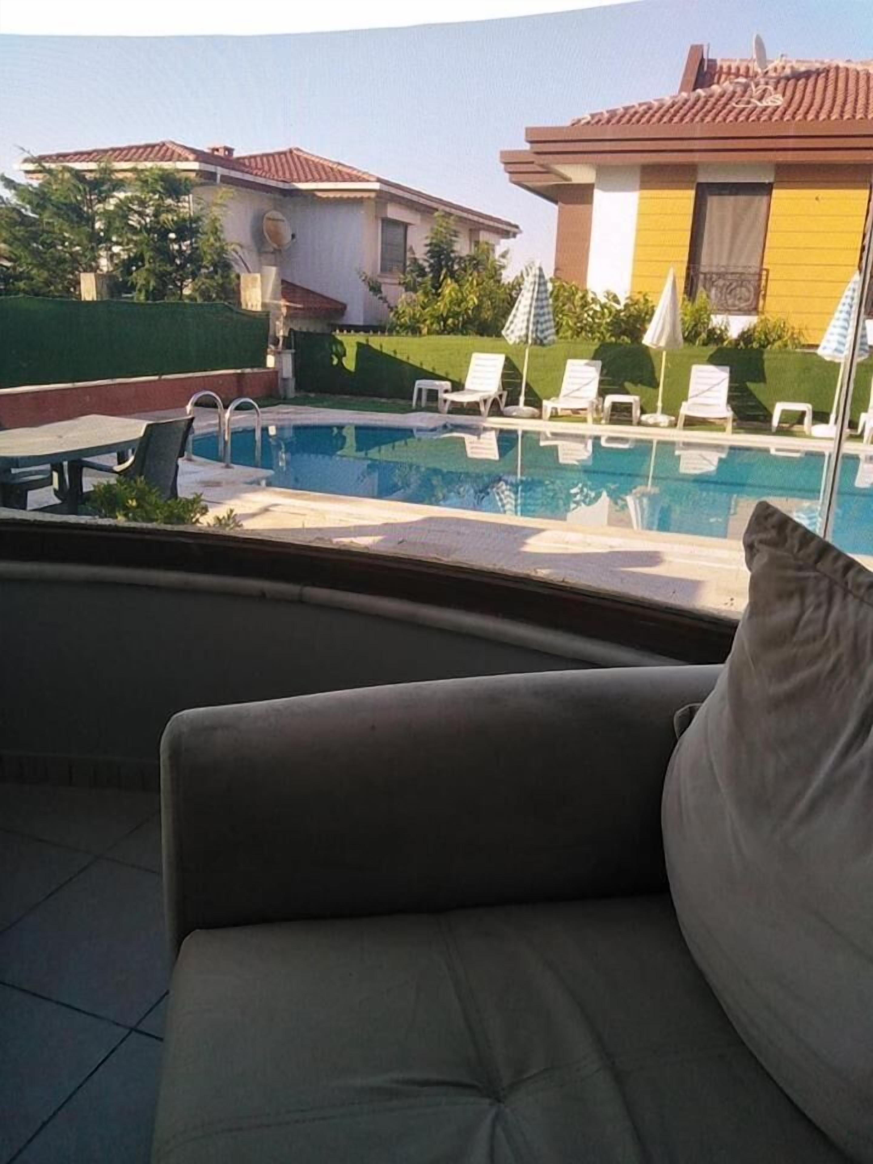 A pool area with lounge chairs and a sofa, a clear view of the pool, and a building with a tiled roof in the background.