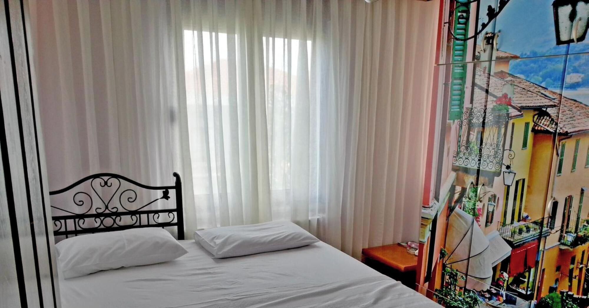 Un dormitori amb un llit, cortines, una taula petita i una paret amb mural.