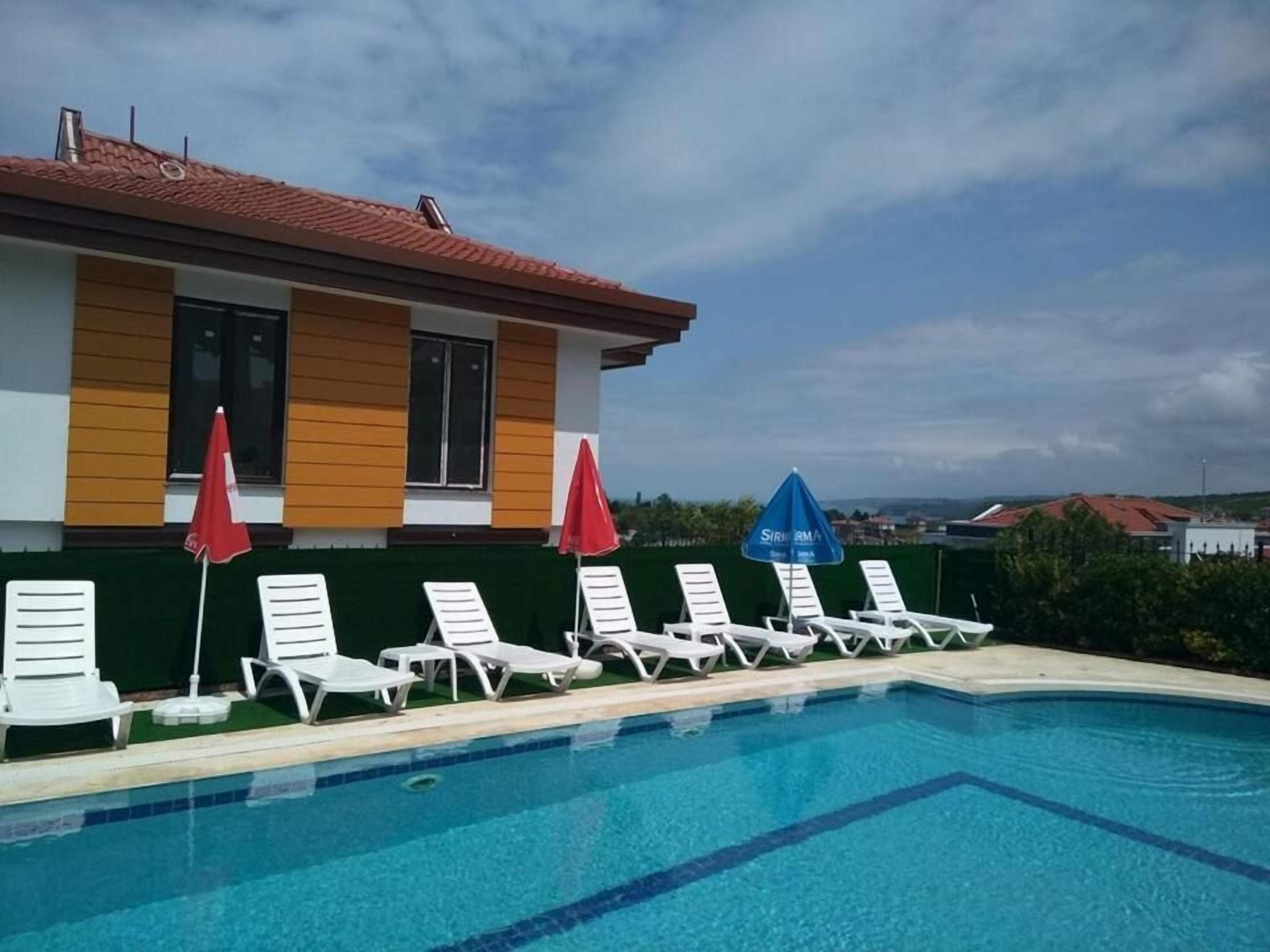 A pool area with white lounge chairs and red umbrellas, a building with a red roof, and a clear sky.