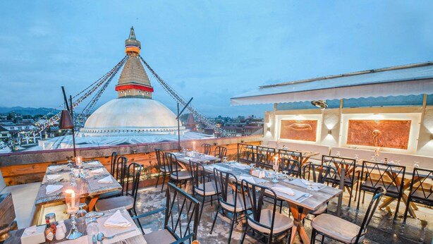 Un restaurante al aire libre con mesas y sillas, un gran edificio blanco con techo puntiagudo al fondo y vistas a la ciudad.
