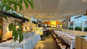 Un restaurante con mesas largas dispuestas para comer, una zona de cocina al fondo y plantas en las paredes.