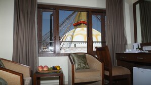 Habitación de hotel con vista a un edificio grande y un puente. Hay una silla, una mesita con fruta y un escritorio con una computadora.