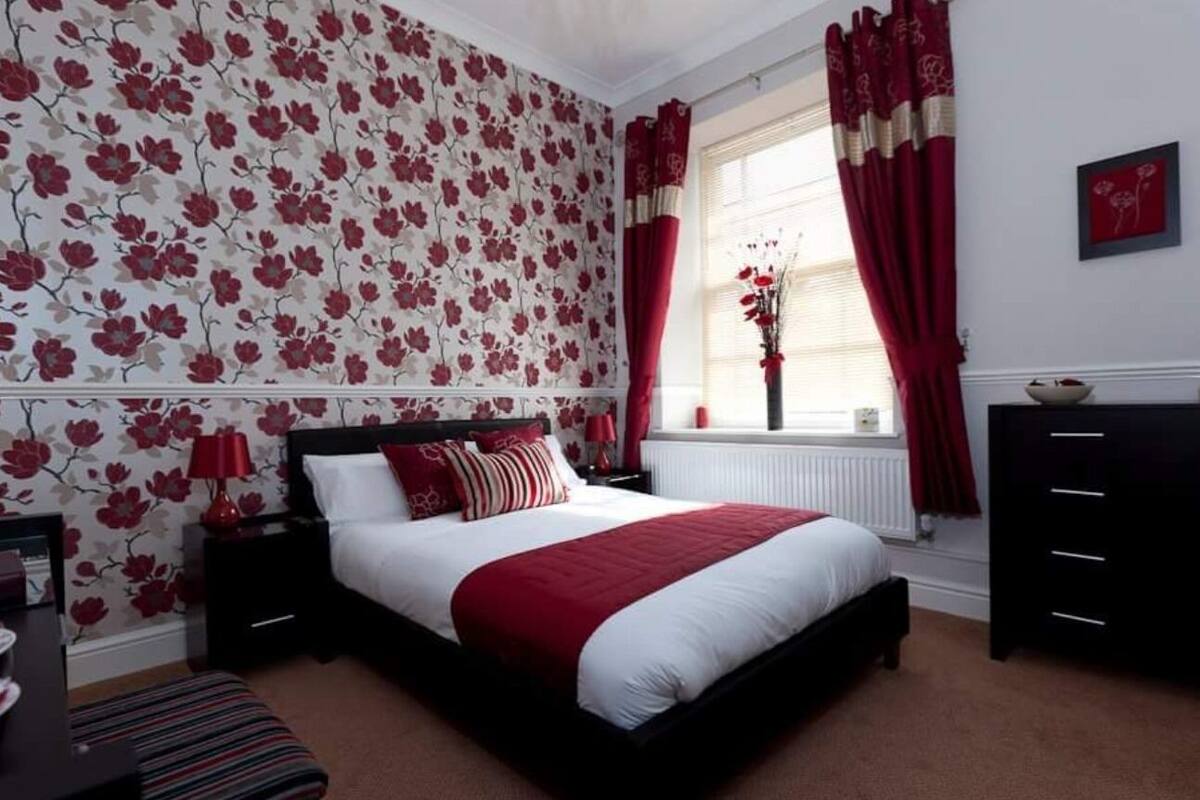 A bedroom with a large bed, a nightstand, a dresser, a window with curtains, and a floral wallpaper.