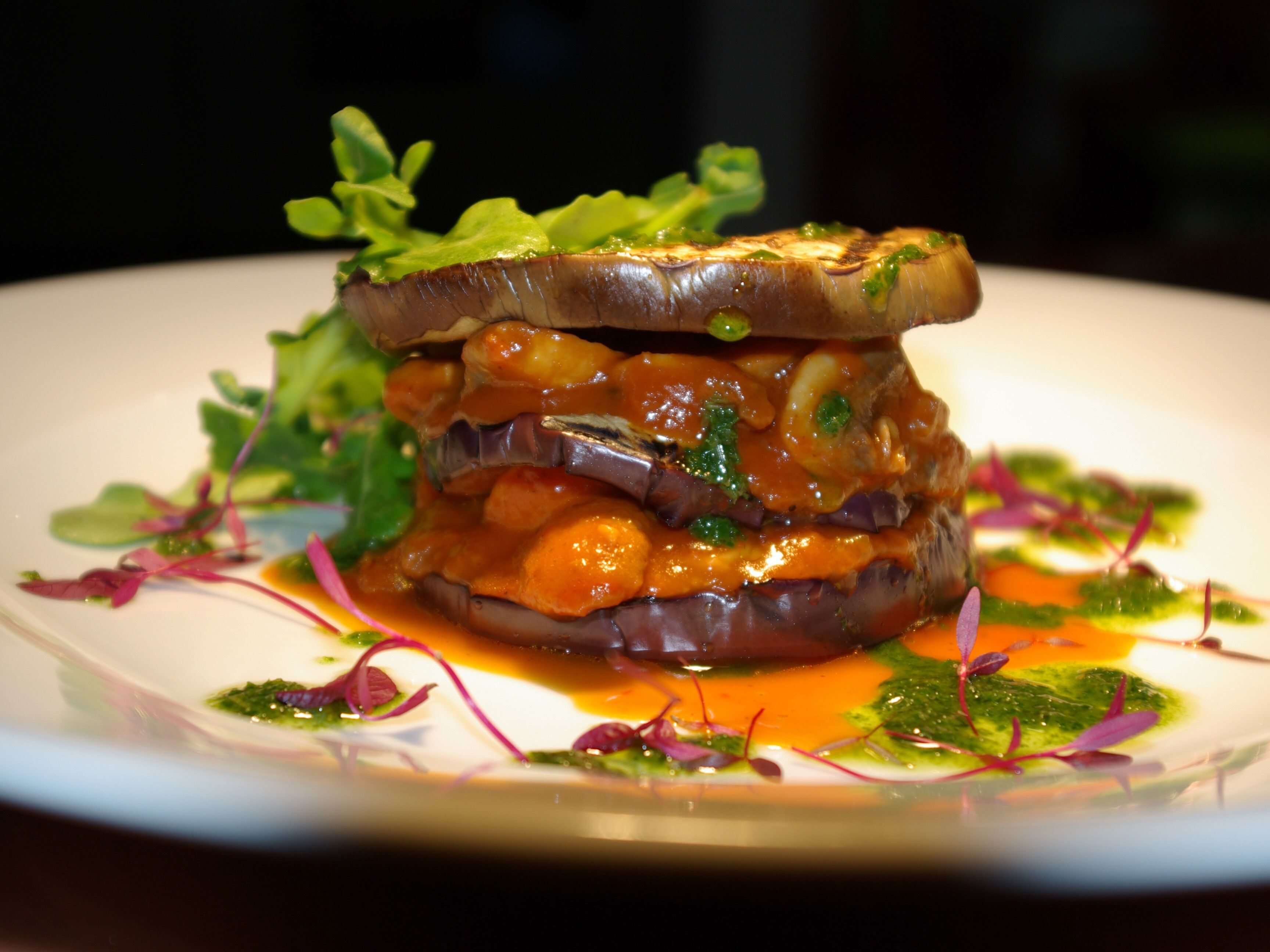A dish with a slice of eggplant and greens on top, surrounded by sauce and greens.