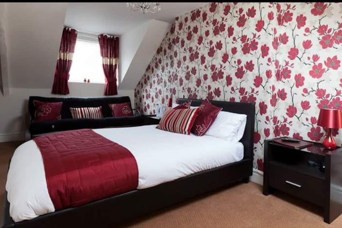 A bedroom with a large bed, dark nightstands, a red lamp, and floral wallpaper.