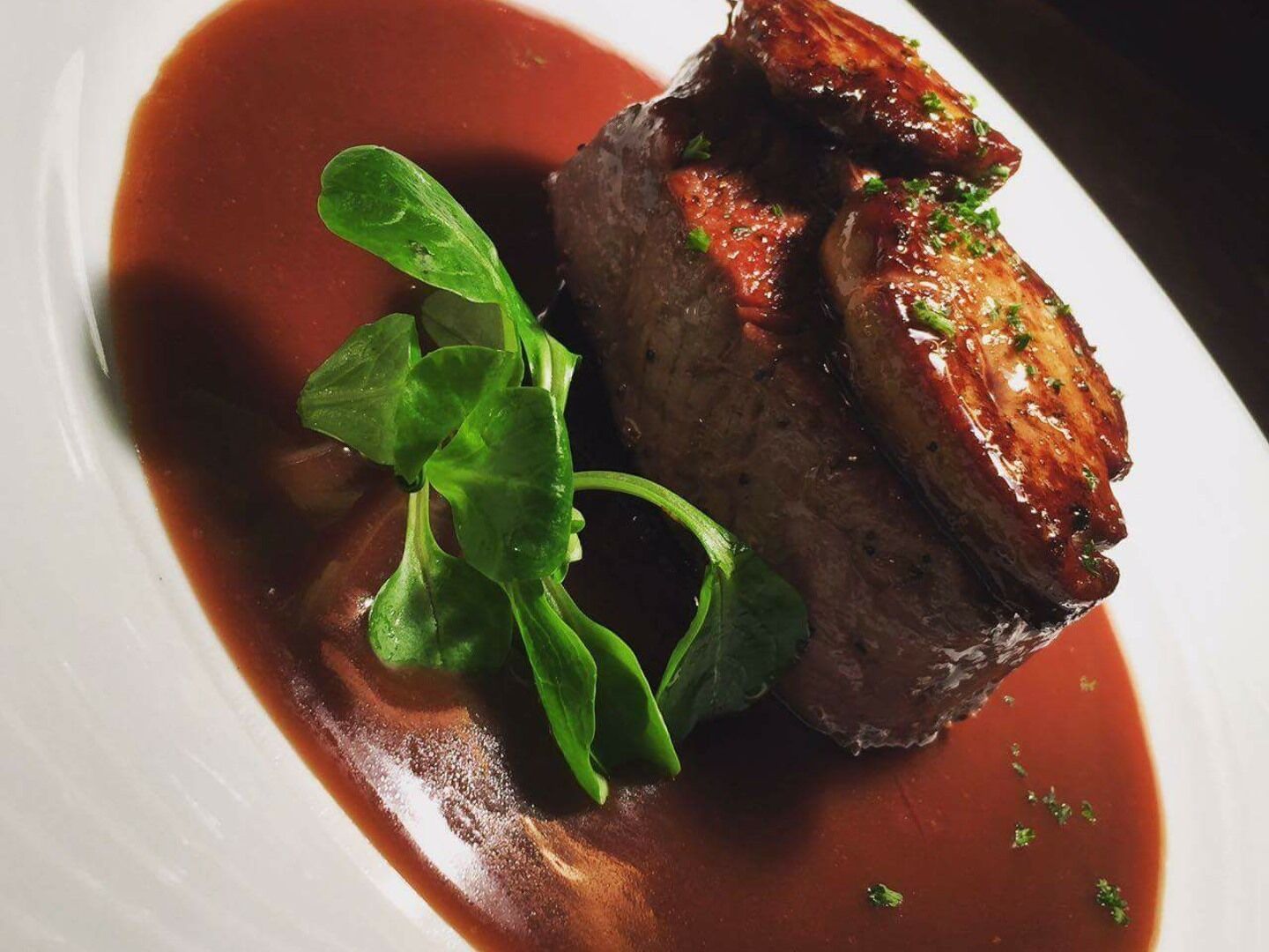 A plate with a piece of meat, possibly steak, garnished with herbs, accompanied by a green leafy vegetable and a red sauce.