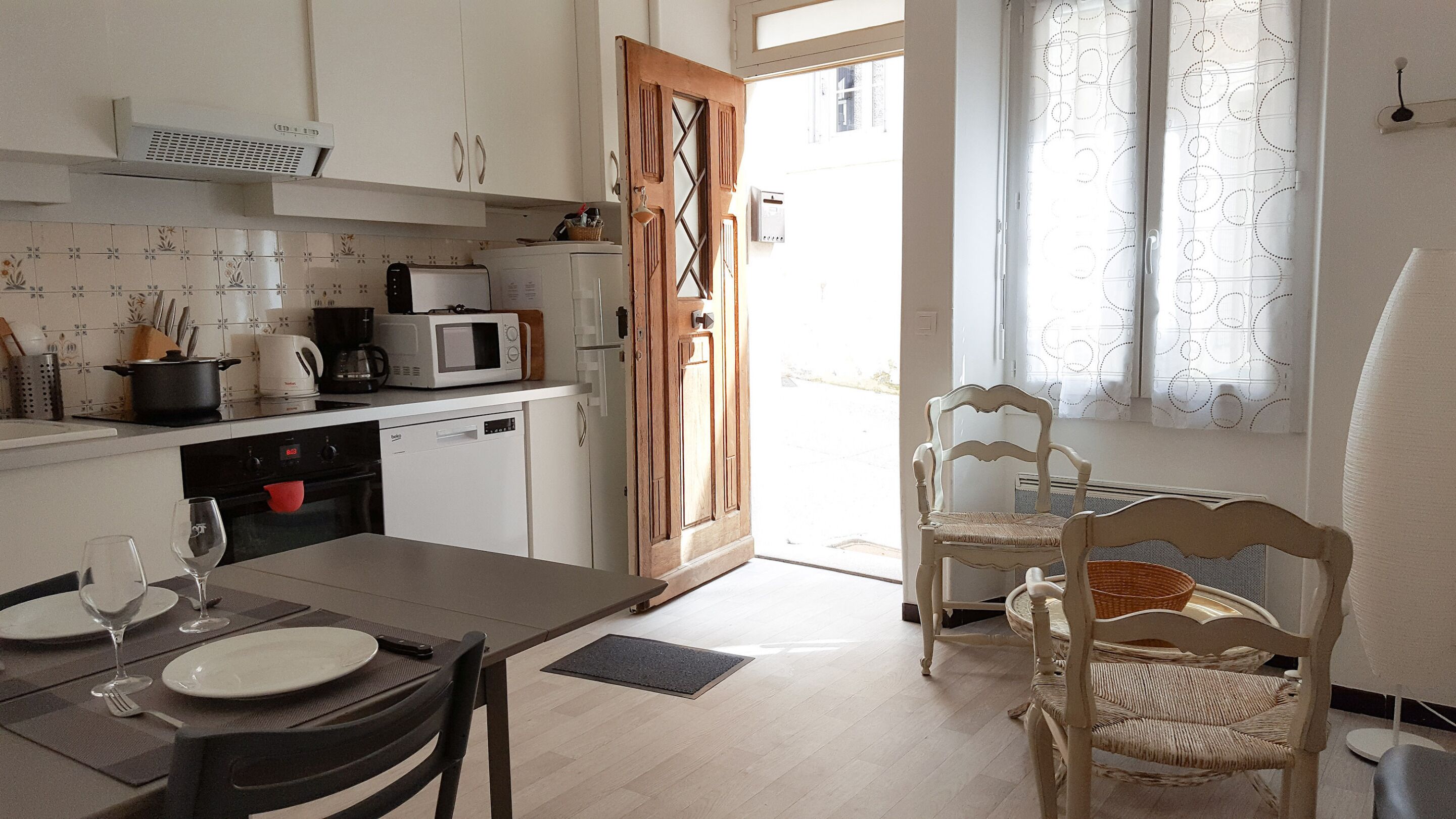Una cocina con gabinetes blancos, microondas, cafetera y tostadora. Hay una mesa de comedor con sillas y una ventana con cortinas.