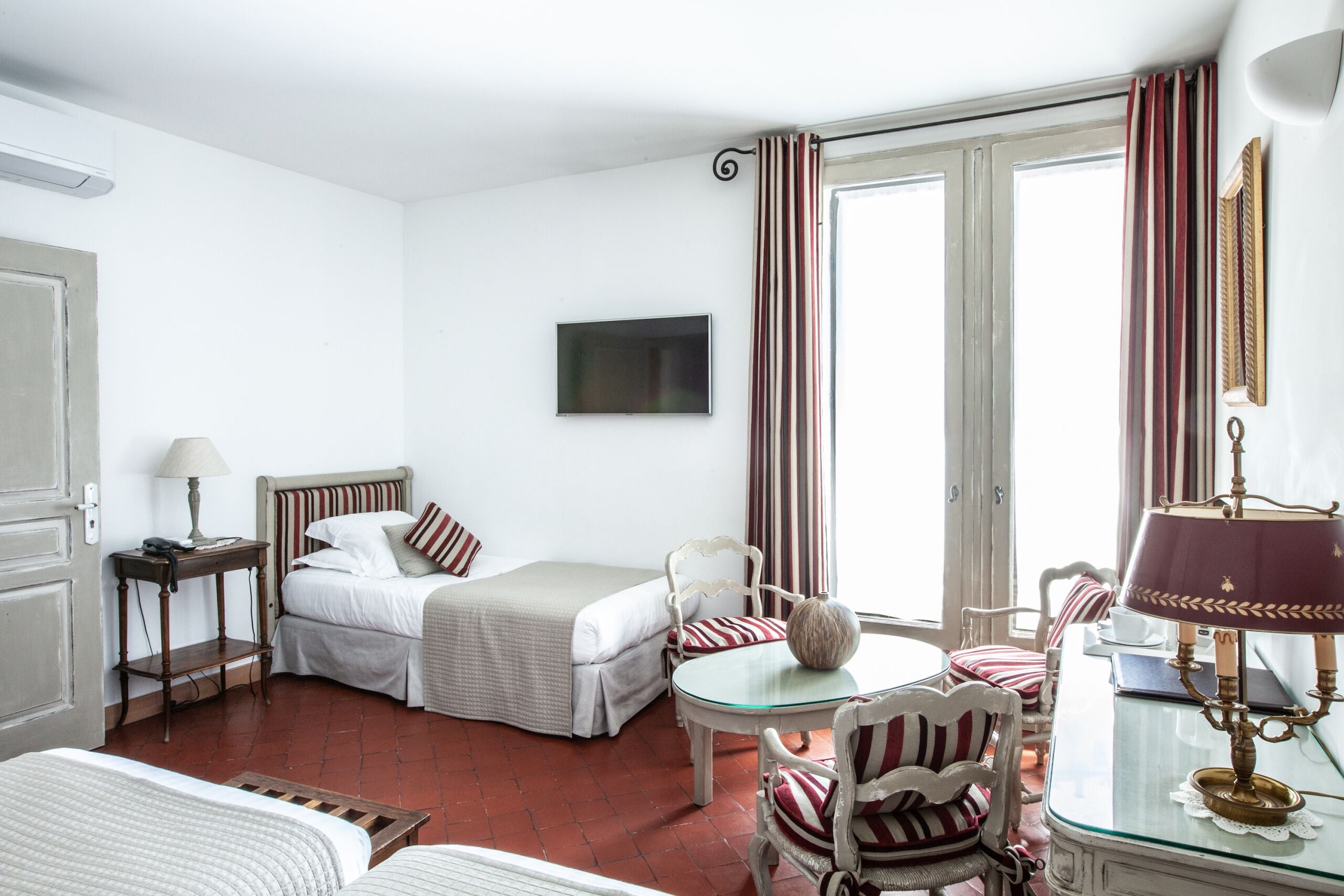 Una habitación de hotel con cama, escritorio con lámpara, silla, televisión y ventana con cortinas.