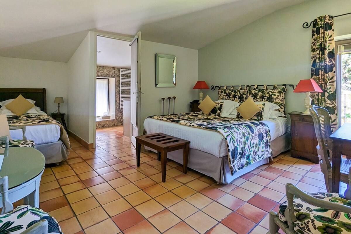 A bedroom with two beds, a small table, and a chair. There is a mirror on the wall and a window with curtains.
