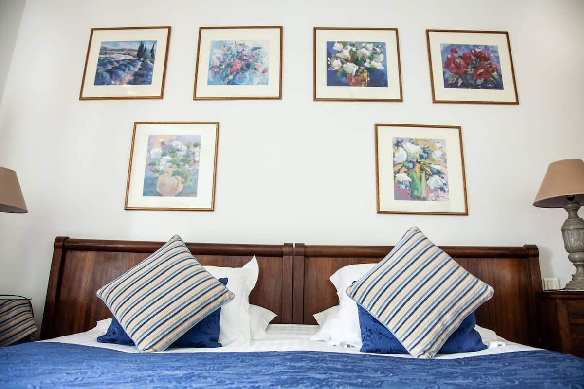A neatly made bed with blue bedding and striped pillows, a wooden headboard, and framed floral paintings on the wall.