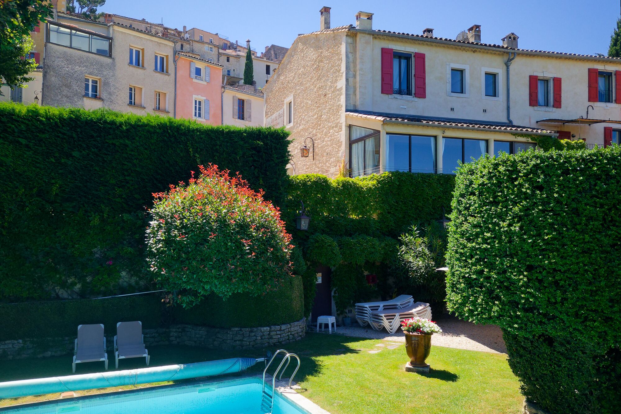 A pool area with lounge chairs, a well-maintained lawn, and a building with stone walls and red shutters in the background.