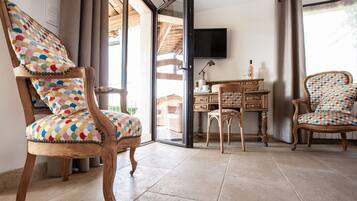 Una habitación con una silla de madera y cojín colorido, un escritorio de madera con televisor, y vistas a través de un ventanal.