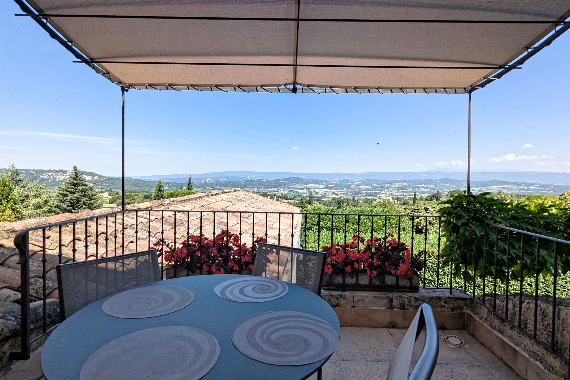 Un espacio de estar al aire libre cubierto, con una mesa y sillas, que ofrece vistas panorámicas a colinas y el mar en la distancia.