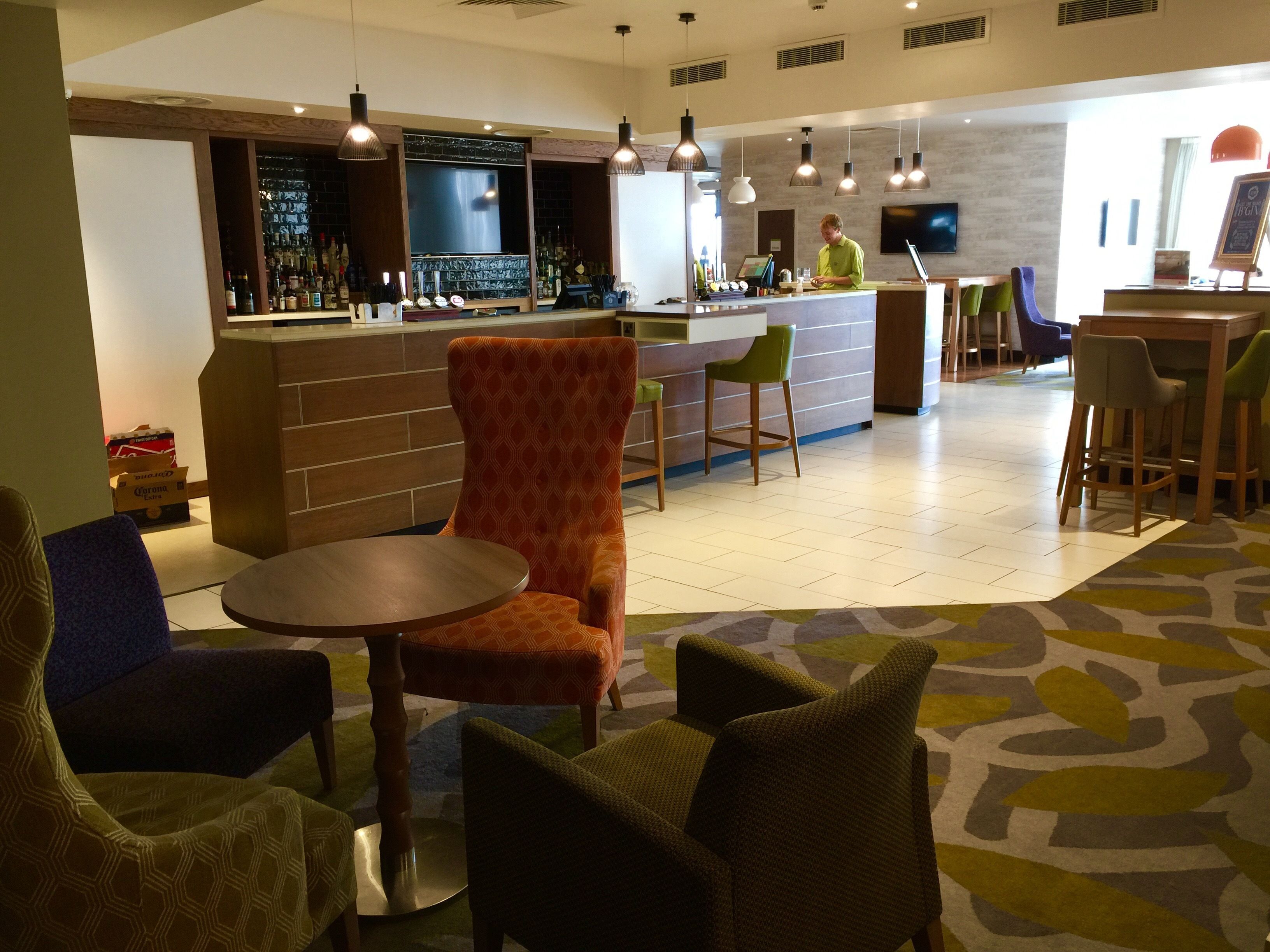 A bar area with a counter, bar stools, and a bartender. There are chairs and a table in the foreground.