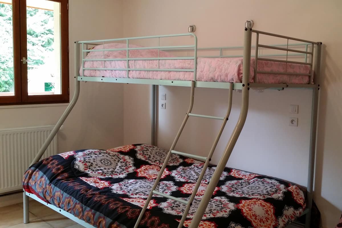 A bunk bed with a patterned bedspread, a window with a view of trees, and a radiator.