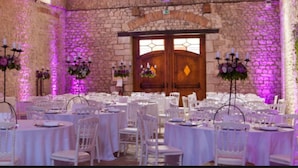 Une salle de banquet avec des tables rondes dressées pour un mariage, des murs en pierre et un éclairage violet.