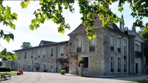 Un grand bâtiment en pierre, doté de nombreuses fenêtres et d’une entrée centrale, entouré d’une allée gravillonnée et de verdure.