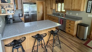A kitchen with wooden cabinets, a stainless steel refrigerator, a granite countertop, and black bar stools. - Beautiful four season chalet in northern Wisconsin Lakeshore (Siren)