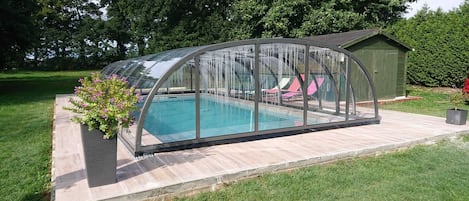 A covered swimming pool with a glass enclosure, surrounded by a wooden deck and a small shed.