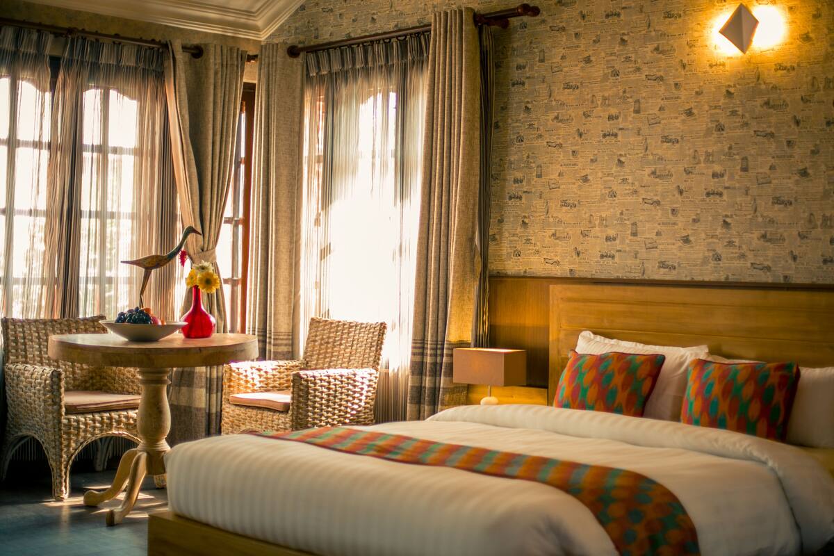 A hotel room with a large bed, a small round table with a vase and fruit, a wicker chair, and a wall with newspaper print.
