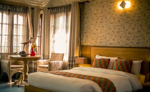 Une chambre d’hôtel avec un grand lit, une petite table ronde ornée d’un vase et de fruits, une chaise en osier et un mur recouvert d’un papier peint à motifs de journaux.