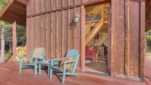 Una cabaña de madera con un porche que tiene dos sillas y una mesa, una escalera visible que conduce al interior, y una pila de leña afuera.