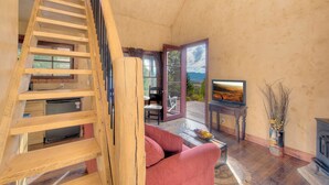 Una escalera de madera que lleva a un balcón con vistas, un sofá rojo, un televisor y un jarrón con ramas.