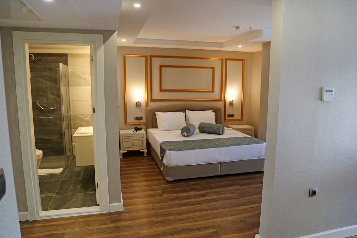 A modern hotel room with a wooden floor, a large bed with white bedding, a shower area, and a wall-mounted light fixture.