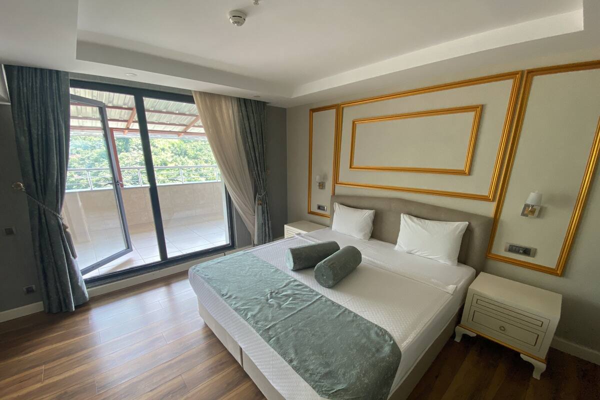 A bedroom with a large bed, a nightstand, a sliding glass door to a balcony, and a ceiling-mounted smoke detector.