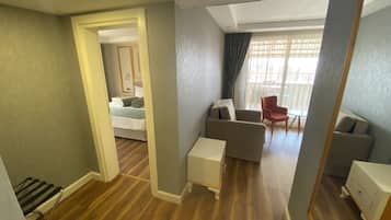 Habitación de hotel con piso de madera, sofá, sillón, mesita y cama con mesita de noche.
