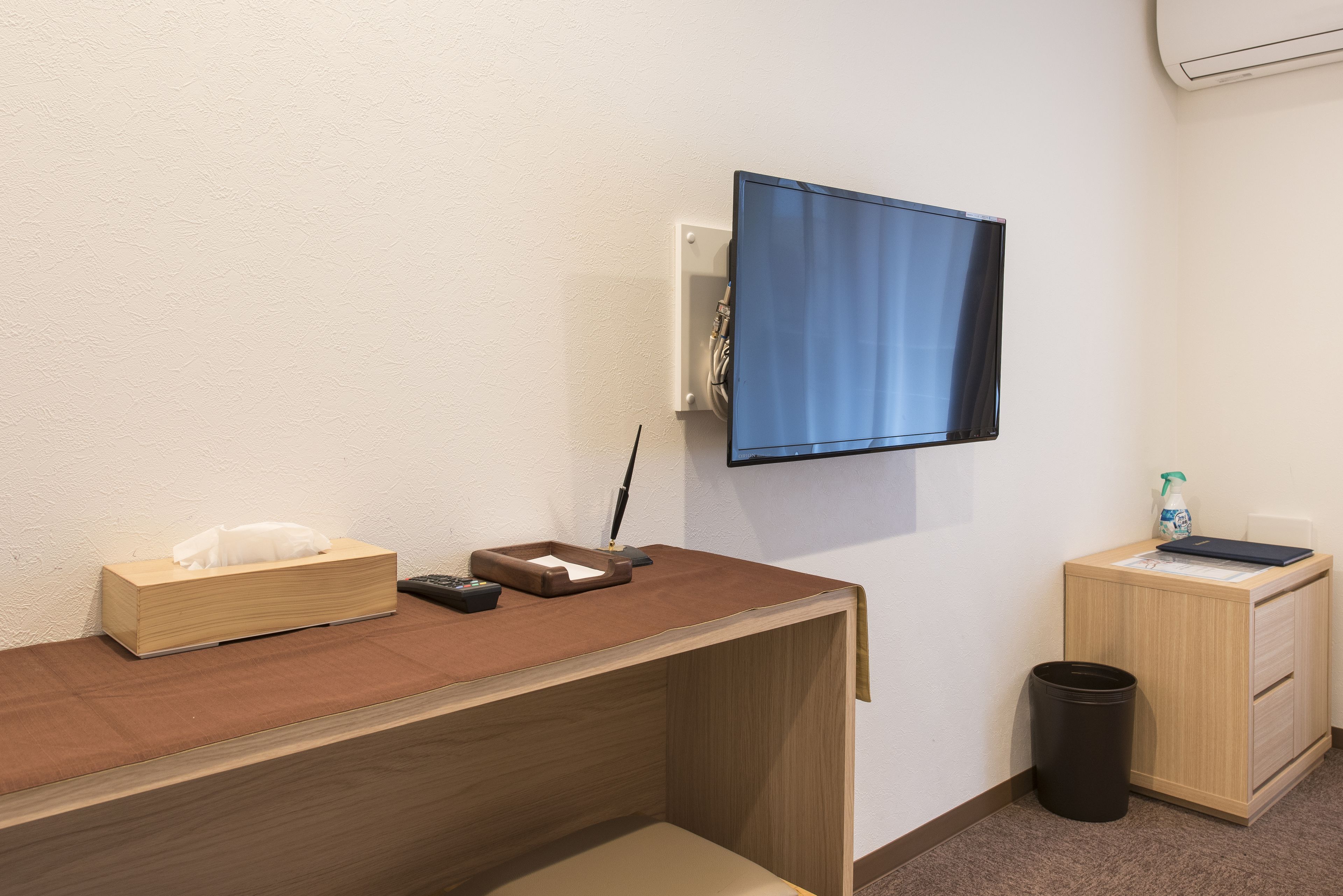 Een hotelkamer met een houten bureau, een aan de muur bevestigde televisie, een kleine prullenbak en een nachtkastje met een lamp en een fles handdesinfecterend middel.