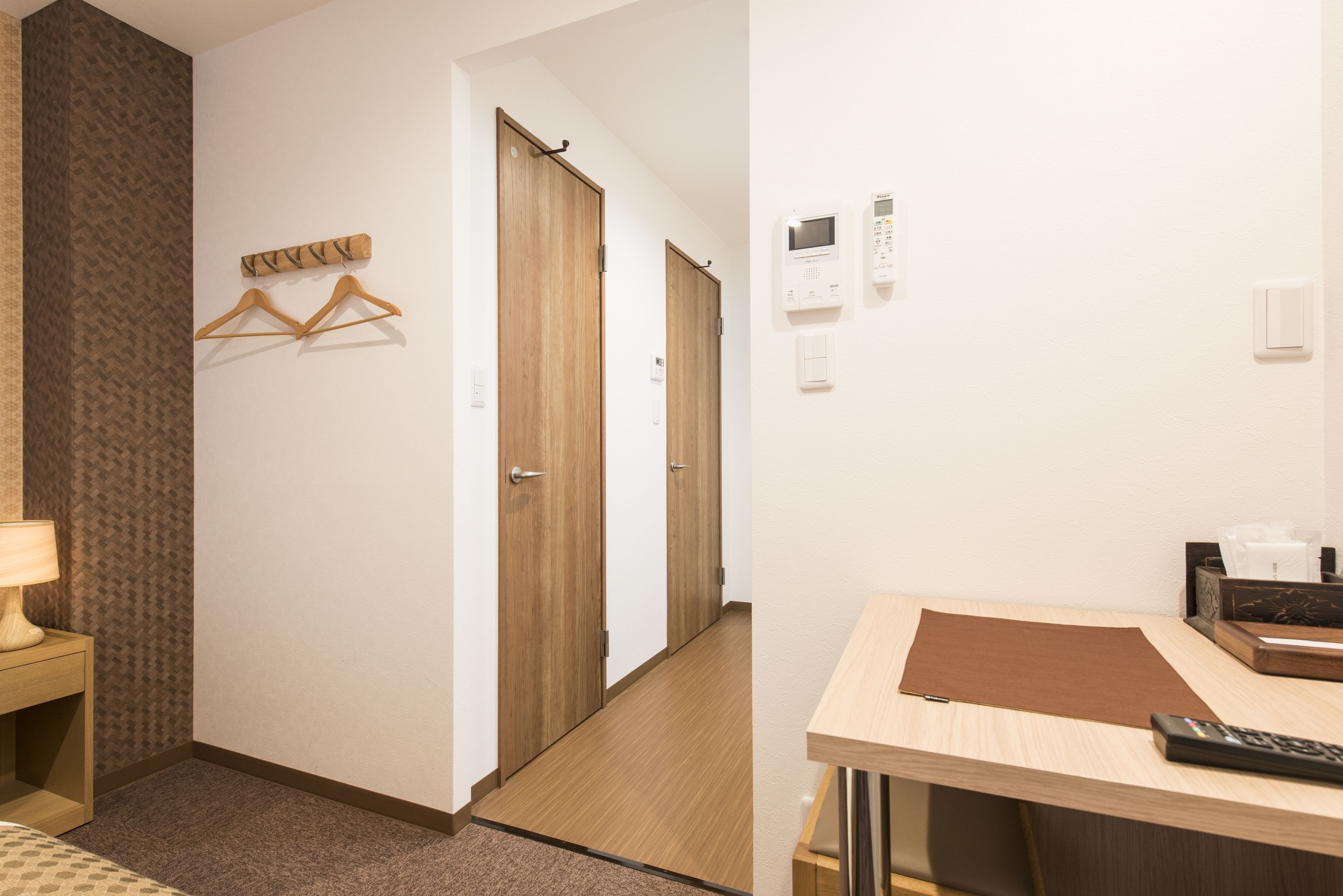 A hotel room with a wooden desk, a lamp, a remote control, and a wall-mounted thermostat.
