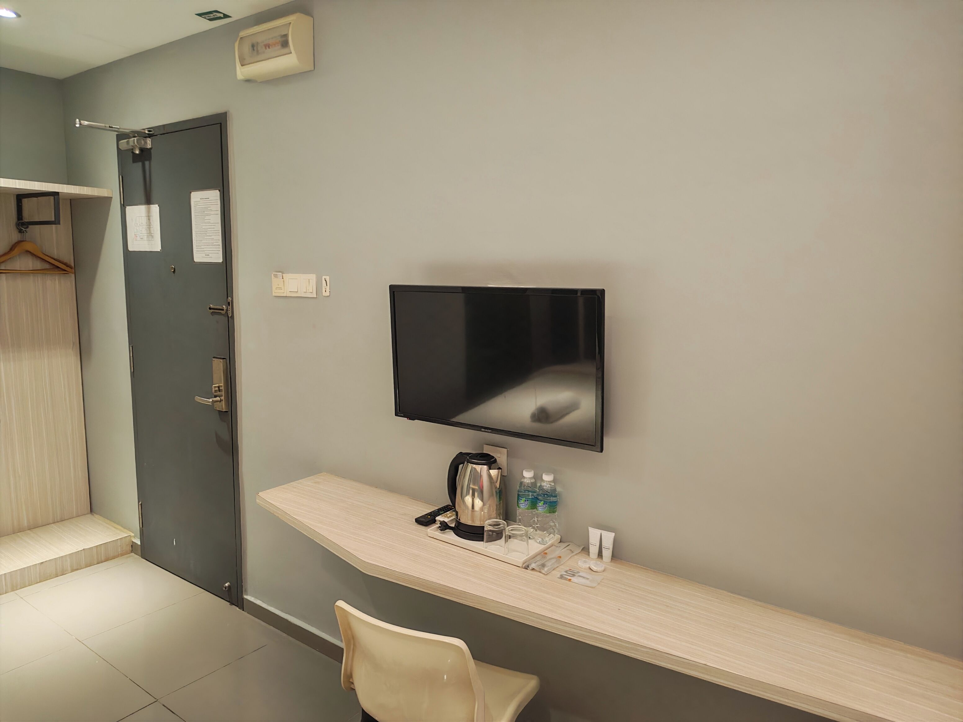 A hotel room with a flat-screen TV mounted on the wall, a desk with a kettle, bottles, and toiletries, and a door with a hanging coat rack.