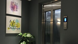 Um elevador moderno com visor digital, uma caixa forte com a etiqueta #95 PERIGO e obras de arte motivacionais nas paredes.
