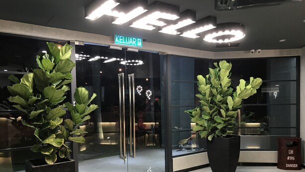 Entrada de hotel com um letreiro 'HELLO', plantas em vasos e um letreiro 'KELUAR'.