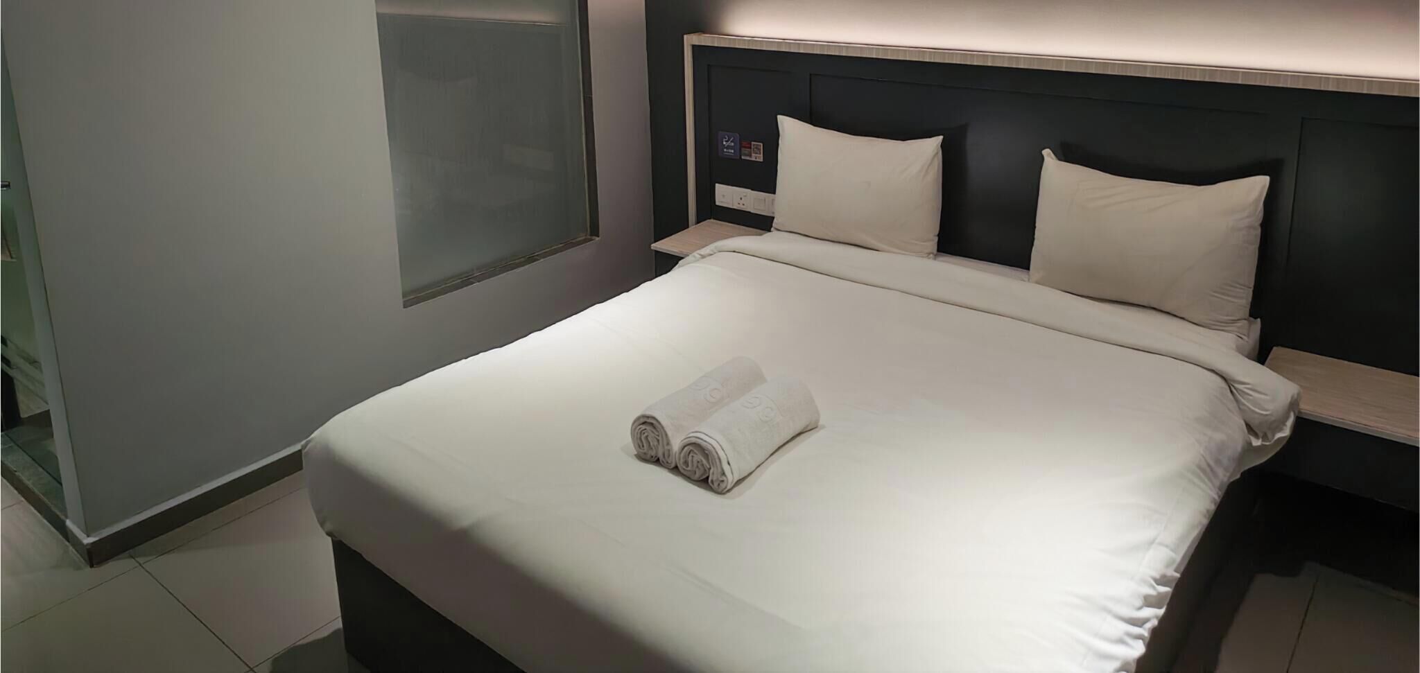A neatly made bed with a rolled white towel, a headboard with a clock, and a bedside table with a power outlet.